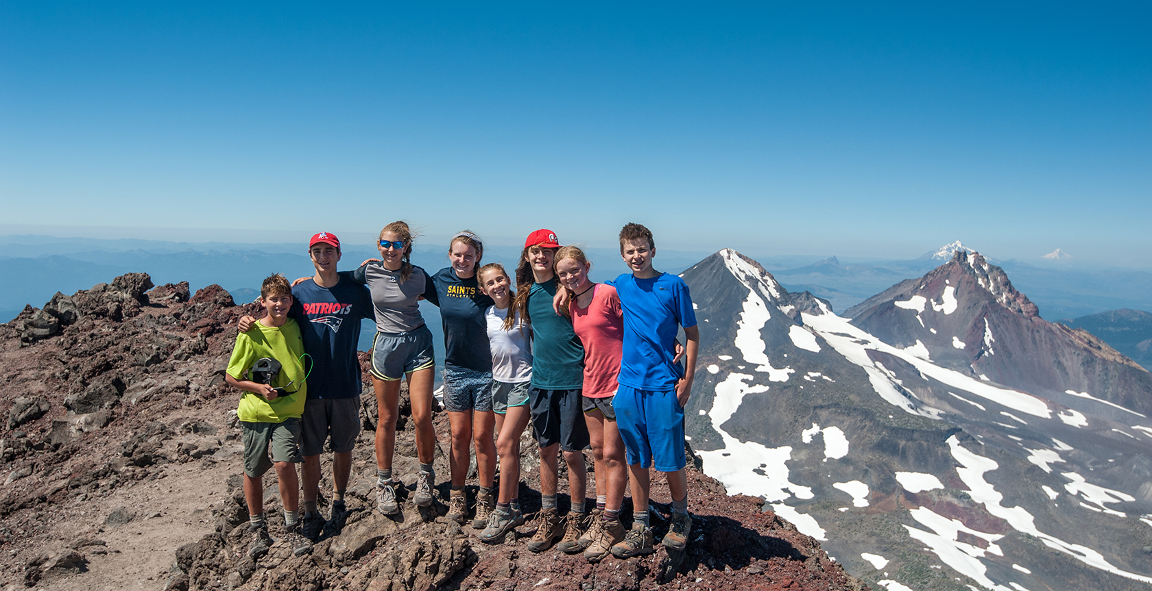 group of kids post hike standing on top of mountain with mountain peaks in background