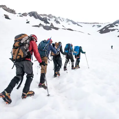 Travelers with backpacks, poles, and shoe spikes, hiking up a snowy mountain