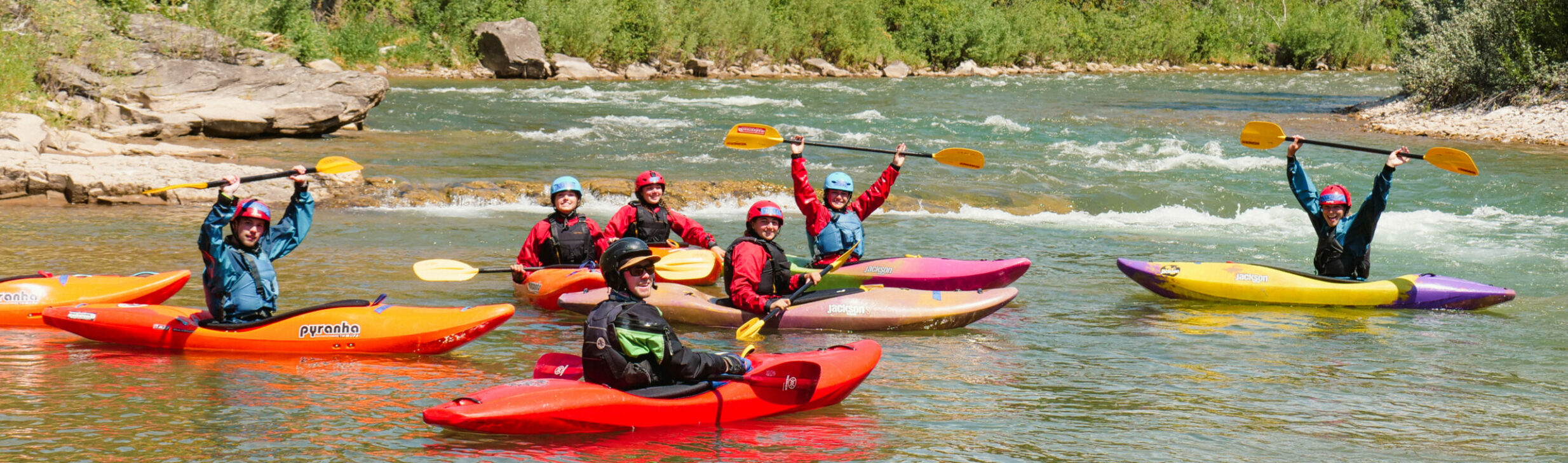 students on a river in whitewater kayaks holding the paddles above their heads