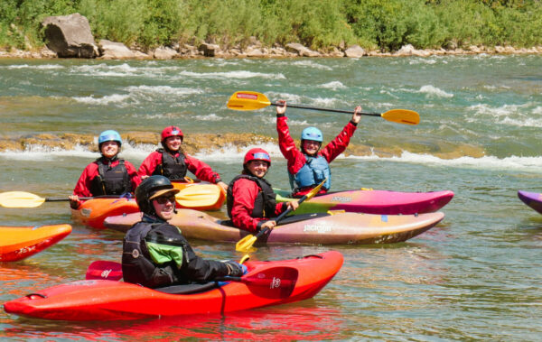 students on a river in whitewater kayaks holding the paddles above their heads