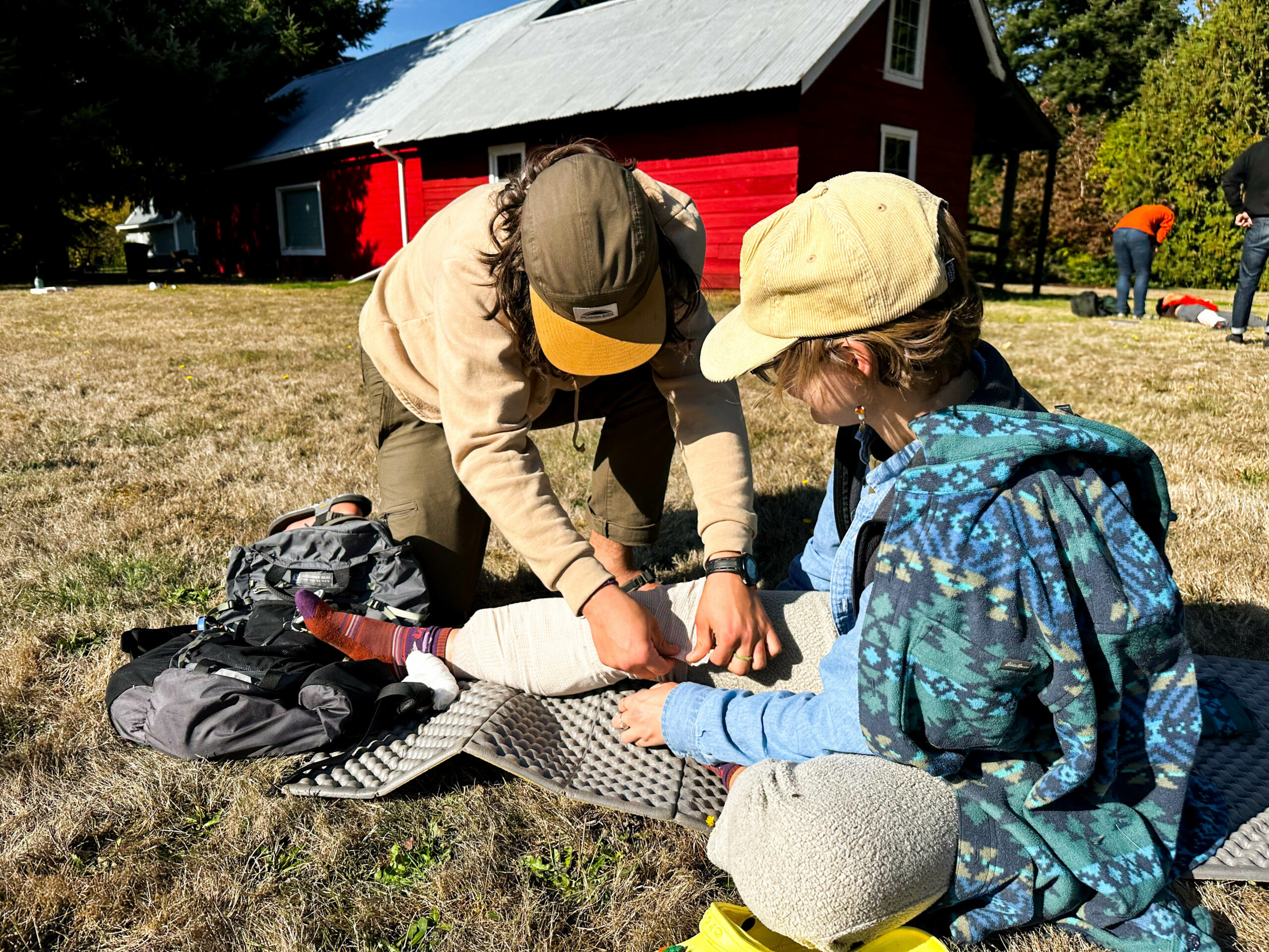 two people sitting in a yard in front of a barn practicing wilderness medicine. One is a patient with a splint on her leg.