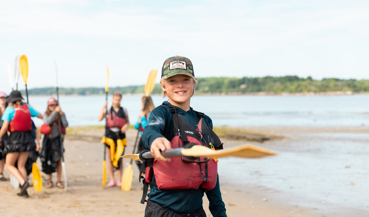 student pointing a kayak paddle at the camera on the shores of a beach