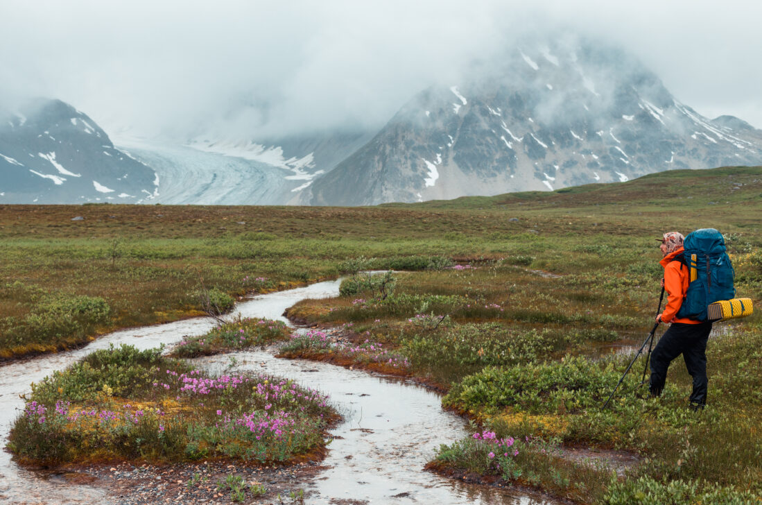 lone backpacker pausing by a river in front of huge cloudy alaska mountains