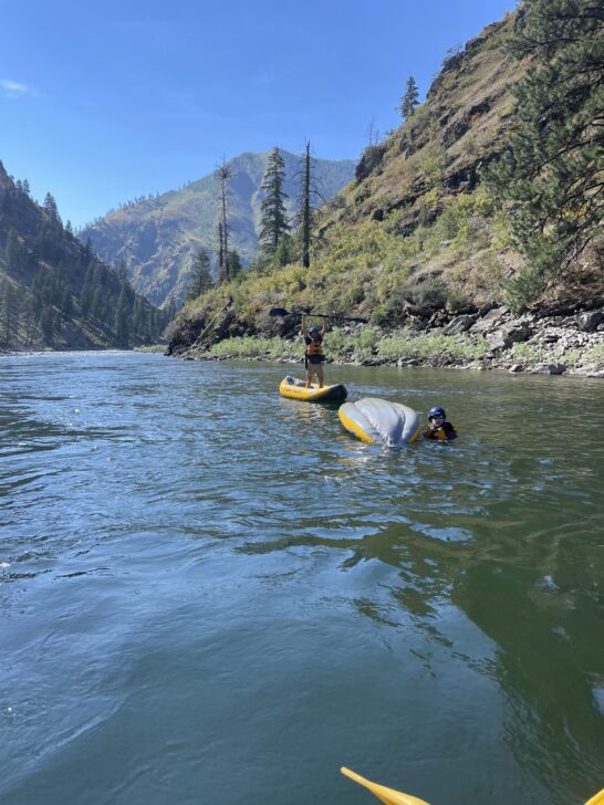 two students in the salmon river, one in an inflatable kayak and one in the river next to a flipped over raft