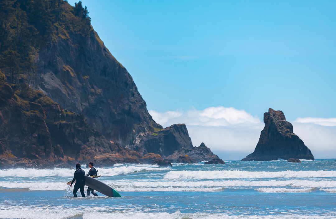 two people in wetsuits carrying surf boards in the water of the oregon coast with rocky cliffs behind them