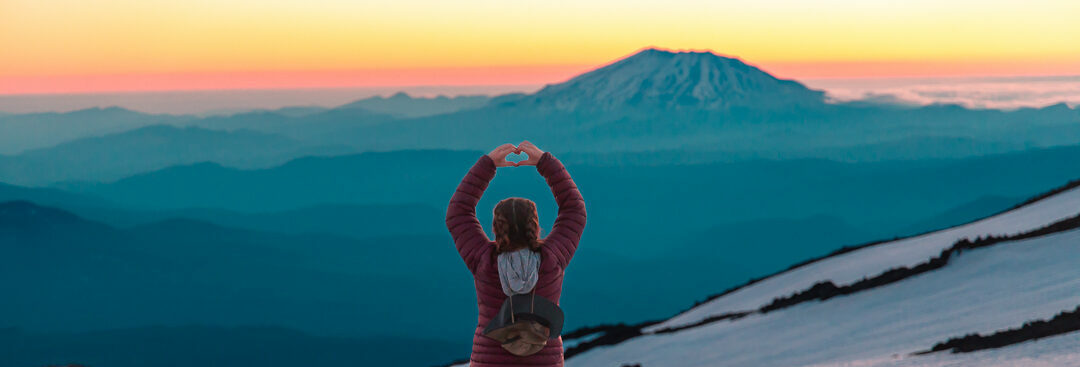 Hiker making a heart with her hands in front of a snowy mountain landscape