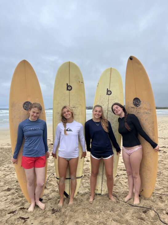 4 girls holding surfboards behind their backs in front of the ocean