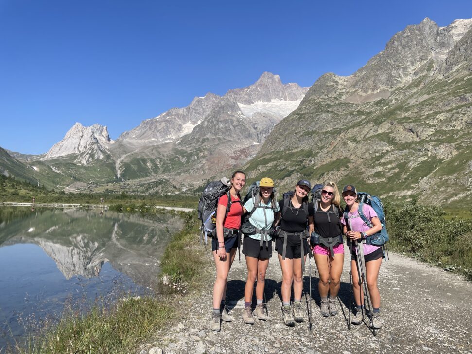 5 students backpacking and posing for a picture in front of an alpine lake and the alps