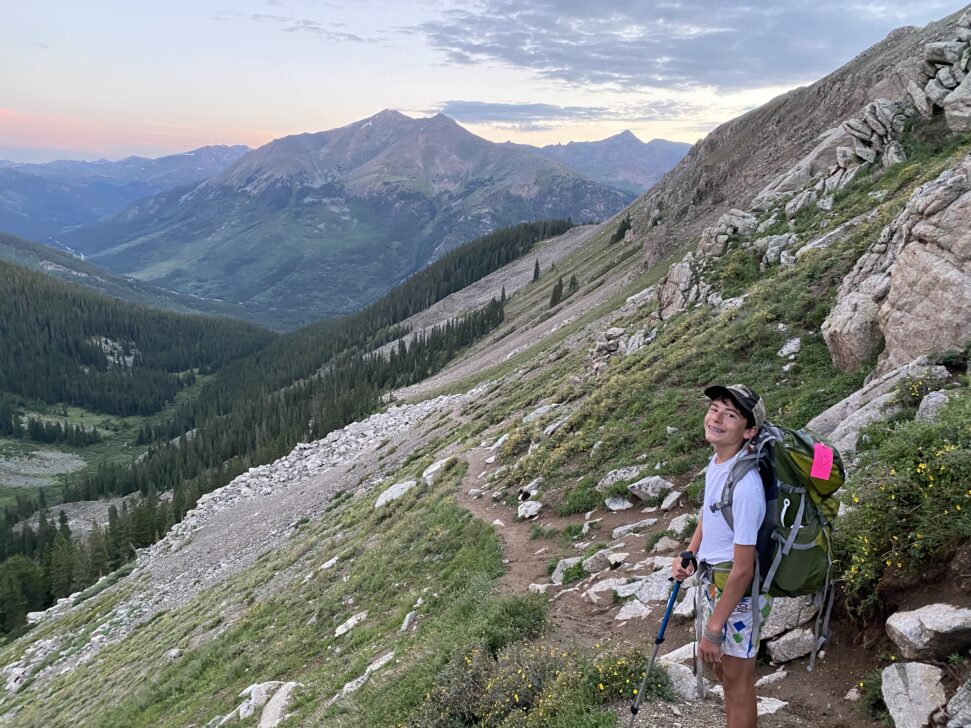 student backpacking in Colorado looking at the camera with the sun setting on mountains behind