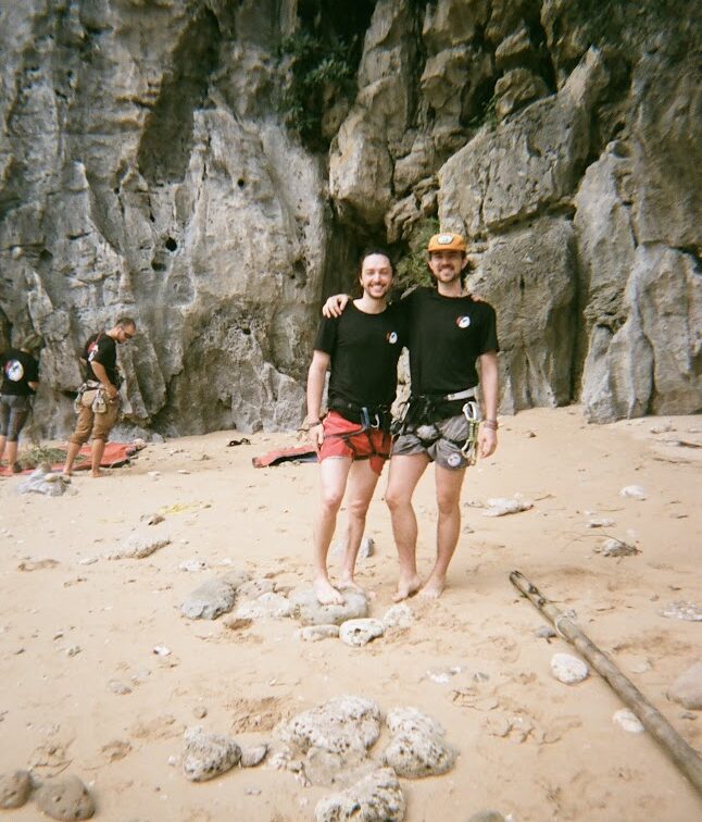 Color film photo of two men in climbing gear with their arms around eachother in front of a climbing wall on a sandy beach.