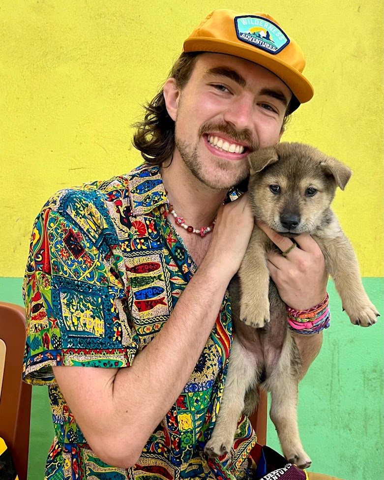 Man in a colorful button-up shirt and yellow baseball hat holding a small puppy next to his face.