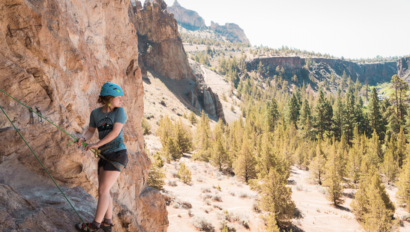 Girl at the top of a rock climbing route looking over her shoulder at vast landscape of pine trees in a valley below her.