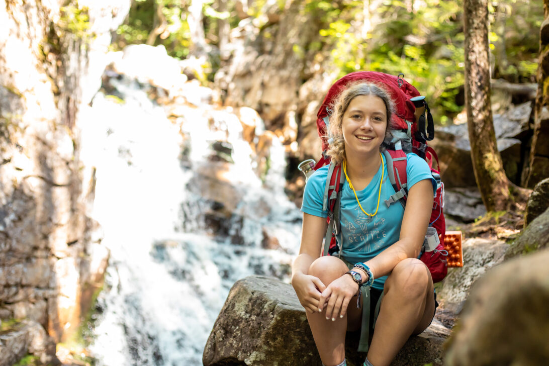 Young girl sitting on a rock with a backpacking pack on in front of a waterfall.