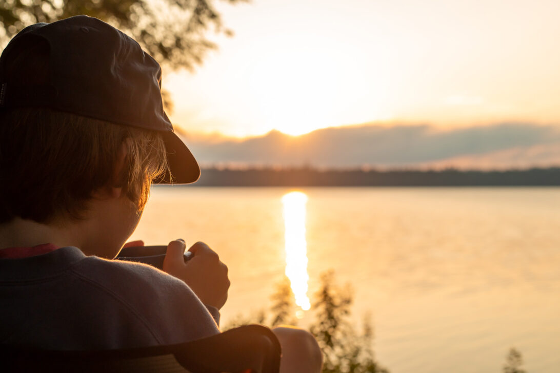 Young person sipping tea looking at an orange sunset over a lake.