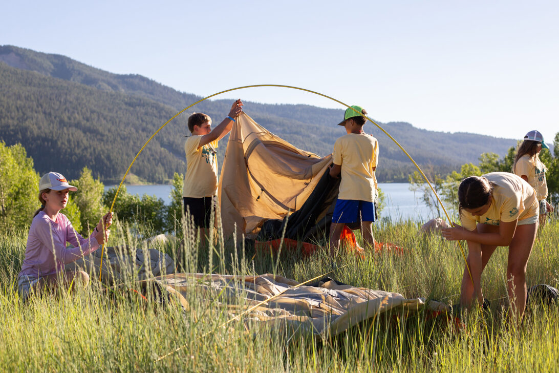 Group of students setting up tents in grasses by a lake.