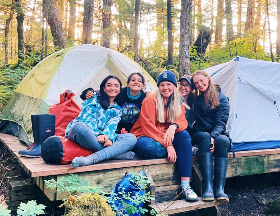 Group of smiling girls sitting in front of tents in the woods.