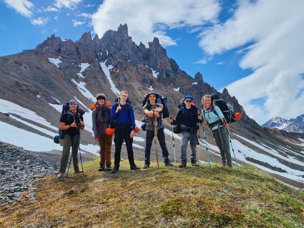 Group of students posing with backpacking packs on in front of a jagged mountain.
