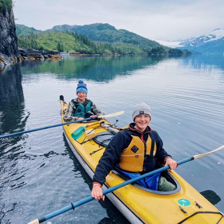 Two kayakers smiling in a yellow kayak on the shore of the ocean in Alaska
