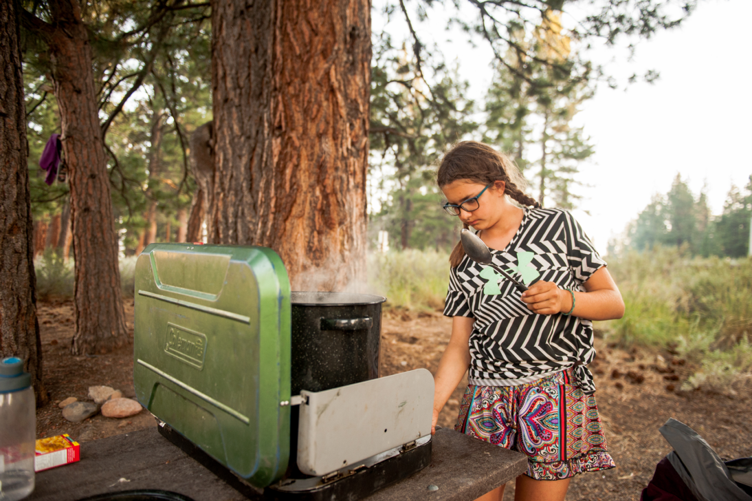 Young camper lighting a stove with a pot of boiling water on a picnic table in the woods.