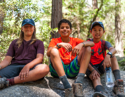 3 young boys sitting on a rock in the woods smiling.
