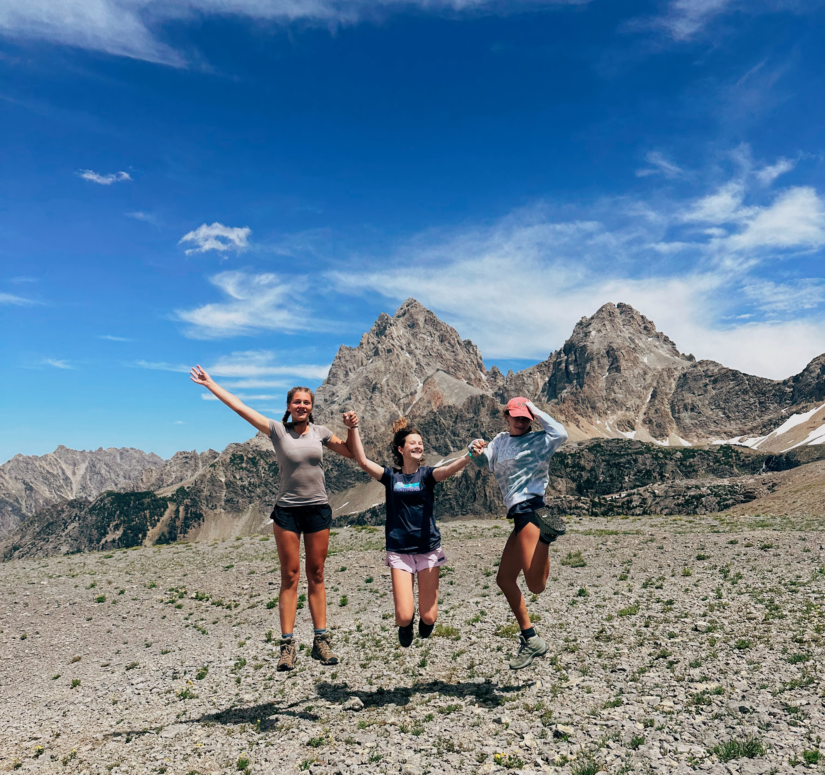 Three girls jumping in the air holding hands in front of a vast mountain on the horizon.