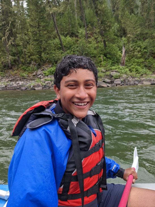 Boy smiling and holding a paddle wearing a PFD on the river.