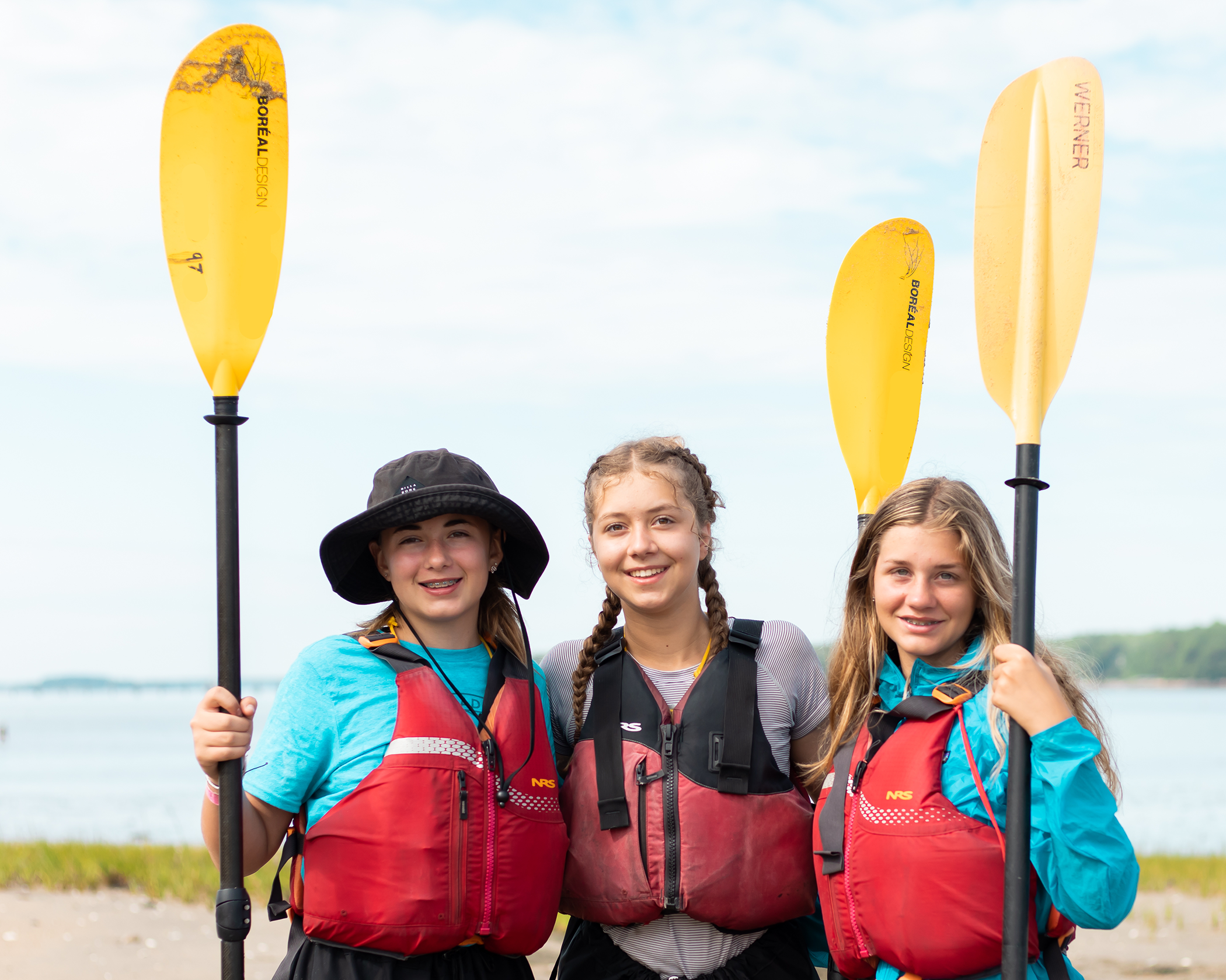 Three girls holding kayak paddles and wearing PFDs in front of the shoreline.
