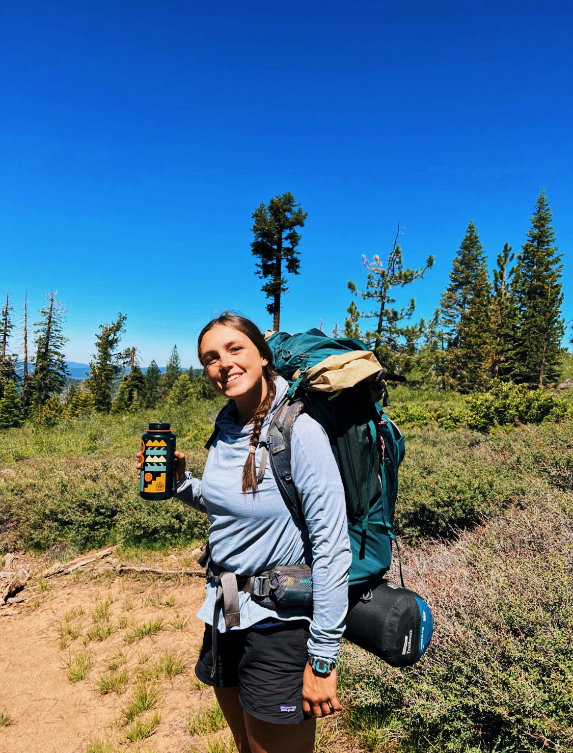 Girl smiling holding a water bottle and wearing a backpacking pack. 